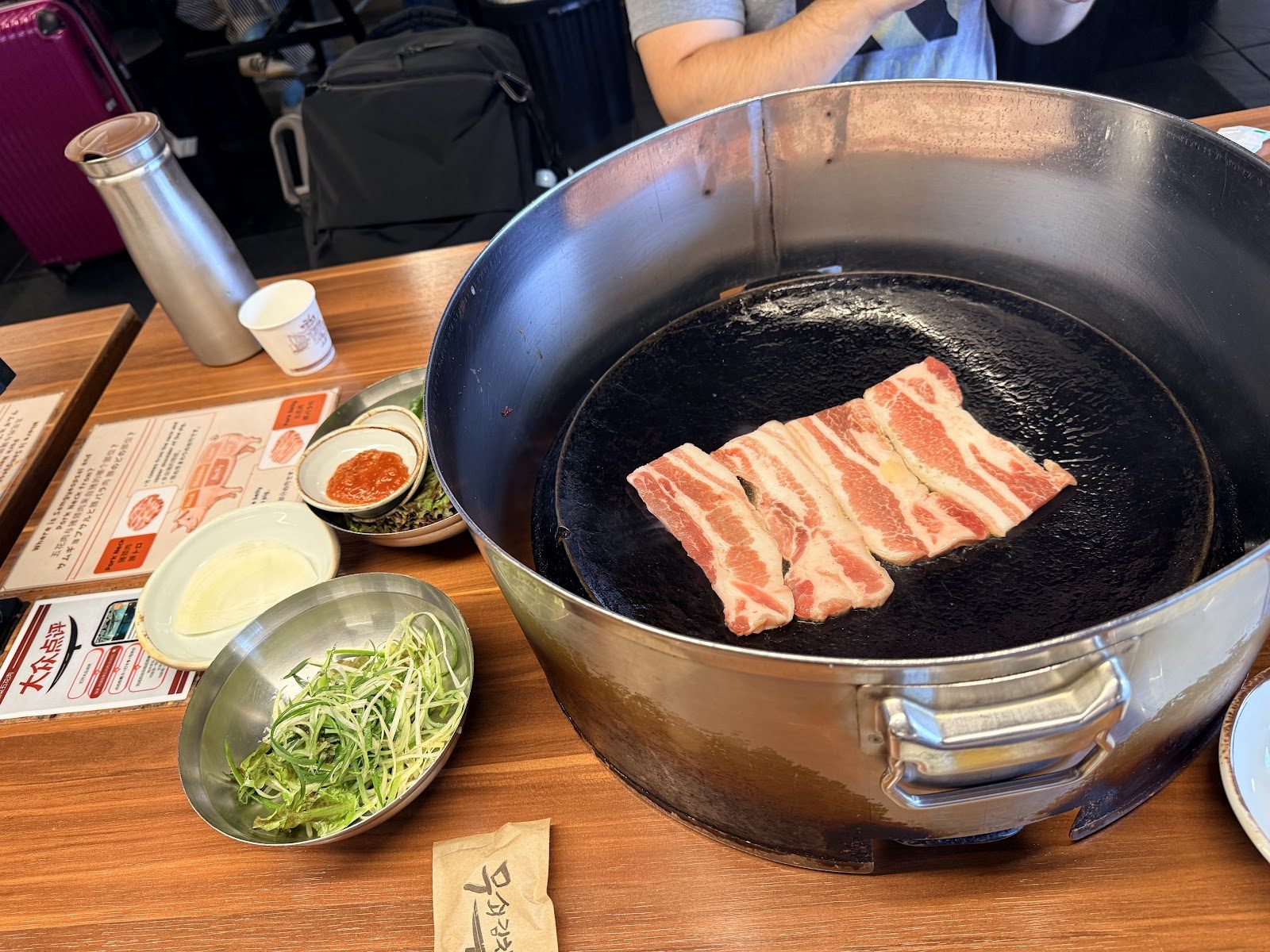 무쇠 뚜껑 모양 불판에 생삼겹살을 막 올린 모습과 옆에 놓인 다국어 안내판, 파채와 쌈장